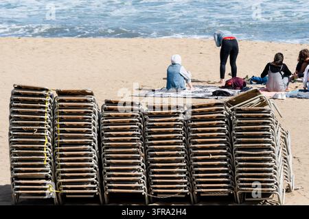GESTAPELTE STRANDSESSLE AM SANDSTRAND, PLAYA DE LA BARCELONETA REPORTAGE, BARCELONA, SPANIEN: Ein großer, ordentlicher Stapel gefalteter gelber und weißer Sonnenliegen steht auf dem Sand, während sich im Hintergrund einige Leute auf Handtüchern am Wasserrand setzen. INFO: Der Strand von Barceloneta ist das berühmteste, lebendigste Meeresziel Barcelonas. Berühmt für seine abschüssigen Sandflächen und die lebendige von Bäumen gesäumte Promenade, verbindet er Sonnenbaden und Wassersport mit geschäftigen Strandbars und Restaurants, Betonpiers und Fitnesszonen. Stockfoto