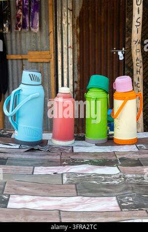 Vier farbenfrohe Plastik-Thermoskannen in Blau, Rot, Grün und Creme, die auf einem gemusterten Tisch vor einer rostigen Wellblechwand an einer Straße aufgereiht sind Stockfoto