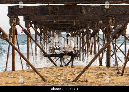 Mann sitzt auf einem Balken unter der verrosteten Struktur eines alten Eisenpiers am Meer, Maritime Region, Lome, Togo Stockfoto