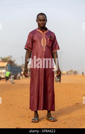 Ein junger Mann trägt ein langes traditionelles dunkelrotes Gewand, während er auf einer staubigen Straße steht, Plateaux-Region, Kpalime, Togo Stockfoto