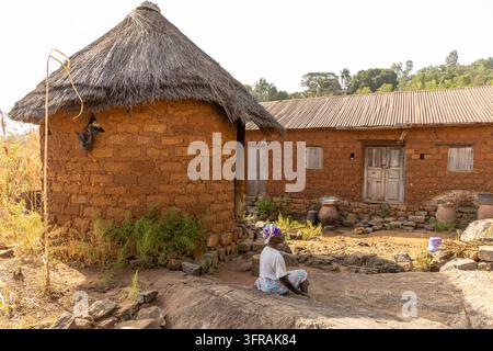 Ein traditionelles rundes Haus aus Lehmziegeln mit einem strohgedeckten kegelförmigen Dach steht in einem ländlichen Komplex, während eine Frau daneben sitzt, Kara-Region, Kabyé-Berg, Togo Stockfoto