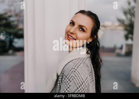 Frau lächelt Porträt einer jungen Brünetten, die eine graue gemusterte Strickjacke trägt und über ihre Schulter blickt, bei einem Tageslichtspaziergang Stockfoto