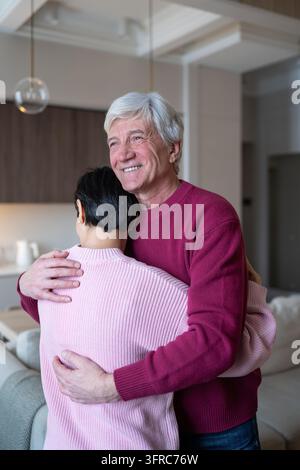 Lächelnder, glücklicher älterer Ehemann umarmt seine liebevolle Ehefrau mit Liebe und Zuneigung im Wohnzimmer. Stockfoto