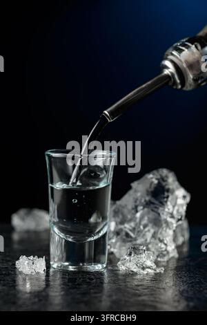 Kristallklarer Wodka oder Gin wird in ein Glas gegossen. Transparentes alkoholisches Getränk mit Eisstücken auf schwarzem Hintergrund. Stockfoto