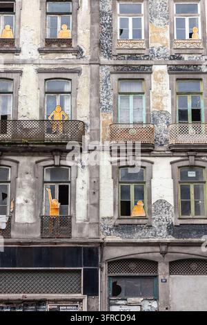 Verlassene Gebäudefassade in Porto, Portugal, mit orangefarbenen Papp-Silhouetten in den Fenstern als urbane Straßenkunstinstallation. Stockfoto