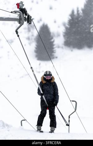 Skifahrer in schwarzer Winterkleidung, Helm und rosa Skibrille fährt einen oberflächlichen Skilift während des Schneefalls, mit schneebedeckten Berghängen und verschwommenem Hintergrund Stockfoto