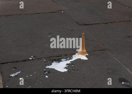 Eine Eiswaffelkegel, der kopfüber auf einem dunklen, strukturierten Asphaltboden gefallen ist, wobei geschmolzenes Vanilleeis eine Verschüttung erzeugt. Stockfoto