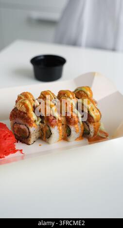 Sushi-Rollen mit bunten Soßen, gebratenen Belägen und eingelegtem Ingwer in einer offenen To-go-Box auf einem weißen Tisch Stockfoto