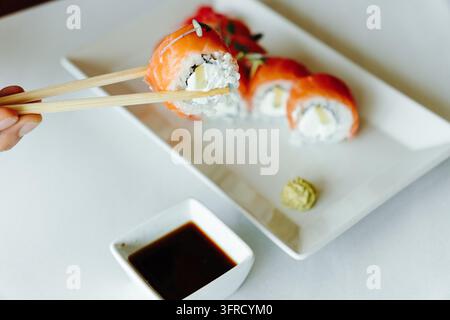Eine Person hält mit hölzernen Essstäbchen eine Lachssushirolle mit Frischkäse und Avocado über einer Schüssel Sojasauce. Stellte japanische Küche dar, resta Stockfoto