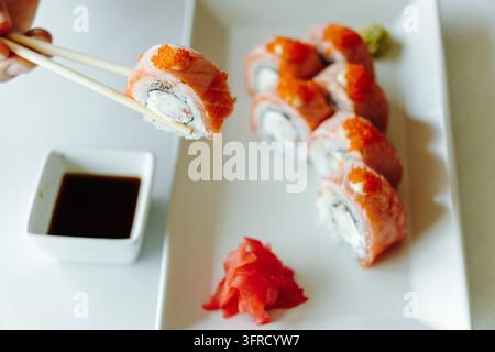Eine Person hält mit Holzessstäbchen eine gebratene Lachs-Sushirolle, die mit orangefarbenem Fischrogen belegt ist, neben einem Teller mit Sushi und Sojasauce. Stellt Japan dar Stockfoto
