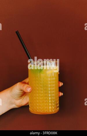 Eine Hand hält ein strukturiertes Glas gefüllt mit einem gelben Zitruscocktail, Minze und einem schwarzen Strohhalm vor einem warmen rötlichen Hintergrund. Vertritt Gastfreundschaft a Stockfoto