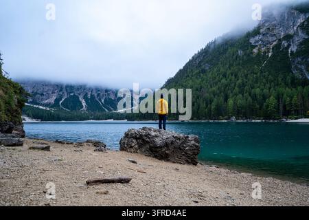 Ein einsamer Wanderer in einer hellgelben Jacke steht auf einem Felsbrocken entlang des Kiesufer eines ruhigen Bergsees. Nebel und Wolkendecke umhüllen den steilen Stockfoto