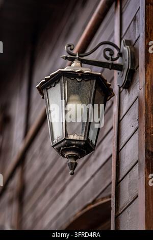 Alte schwarze schmiedeeiserne Wandlaterne mit kunstvollem geschwungenem Brackett, staubigen Glasscheiben und einer mattierten Glühbirne, montiert an einer verwitterten dunklen Holzwand Stockfoto