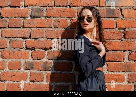 Bezauberndes Mädchen in einem Vintage-Tweed-Mantel, das ein Glas hält und wegschaut. Mädchen mit Sonnenbrille in der Nähe einer weißen Ziegelwand. Warschau Stockfoto