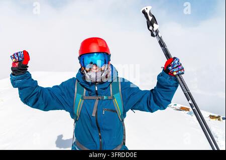 Junger Mann steht im Schnee mit erhobenen Händen und Skistöcken, Freude und Glück im Gesicht des Mannes, der den Berg bestiegen hat Stockfoto