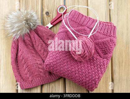 Oberansicht von rosa handgestrickter Winterkleidung mit Pomponmütze, gefaltetem Pullover, Garnballen und Rundstricknadeln, die auf einer Holzoberfläche angeordnet sind Stockfoto