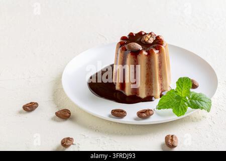 Italienisches Dessert Panna Cotta mit Schokoladensoße und dekoriert mit Kaffeebohnen wird auf einem weißen Teller mit einem Minzgezweig serviert Stockfoto