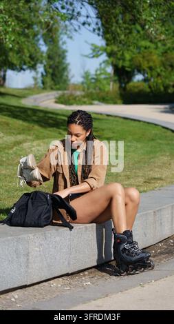 Frau sitzt auf einer Kante und verstaut Sneaker, nachdem sie Rollerblades angezogen hat, in einem sonnigen Park Stockfoto