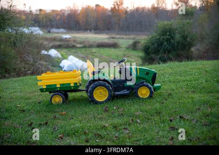 Nostalgisches Familienbild von Kindern, die auf einem Spielzeugtraktor in einer idyllischen Naturkulisse mit grünem Gras und landwirtschaftlichen Feldern fahren Stockfoto