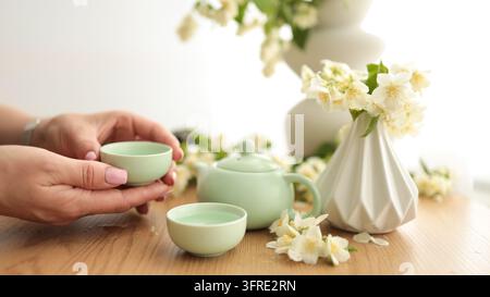 Winkeldetailaufnahme von Händen, die eine pastellfarbene Keramik-Teetasse neben einer Vase mit blühenden weißen Blumen halten. Stockfoto