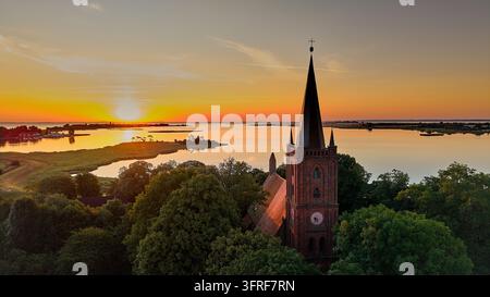 Ein atemberaubender Luftbildaufnahme zeigt eine historische rote Backsteinkirche mit ihrem markanten Turm und Glockenturm, die in dem warmen, goldenen Licht eines **stun** gebadet ist Stockfoto