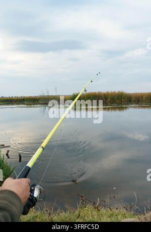 Ich-Perspektive beim Freizeitfischen mit einer Angelrute über einem ruhigen See, der von Schilf und Bäumen gesäumt ist. Stockfoto
