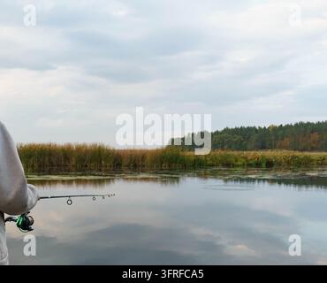 Ich-Perspektive beim Freizeitfischen mit einer Angelrute über einem ruhigen See, der von Schilf und Bäumen gesäumt ist. Stockfoto