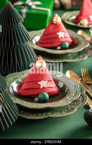 Grüne festliche Tischdeko mit Kugeln geschmückt. Dreifach gefaltete rote Serviette auf dem Teller. Elegantes Feiertagskonzept-Layout. Stockfoto