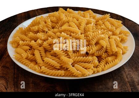 Nahaufnahme von ungekochter Fusilli-Pasta, die auf einem weißen Teller auf dunklem Holztisch gestapelt ist, warmes Licht hebt die Spiraltextur und Goldgelbes hervor Stockfoto