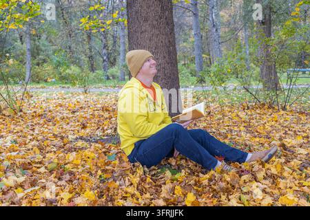 Mann sitzt unter einem Baum auf gelben Blättern und liest ein Buch im Herbstwald Stockfoto