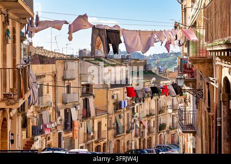 Wäscheleinen mit trocknender Wäsche erstrecken sich zwischen Balkonen einer Wohnstraße, die mit alten Steinhäusern gesäumt ist, in Caltagirone Sizilien, Italien Stockfoto