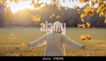 Lächelnde Frau steht mit offenen Armen zwischen vielen fallenden Ahornblättern im warmen Licht der Goldenen Stunde. Breite Herbstkomposition, die Freude, Freiheit, Fürsorge ausdrückt Stockfoto