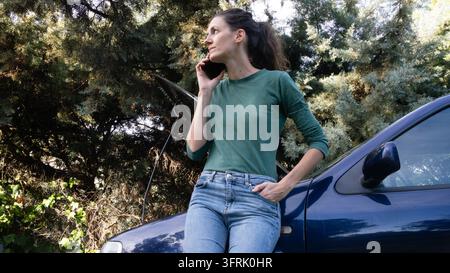 Frau, die einen Telefonanruf tätigt, während sie neben einem liegengebliebenen Auto mit geöffneter Motorhaube steht Stockfoto