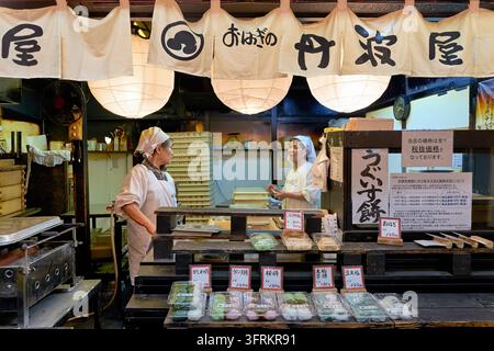 Zwei Frauen mit weißen Kopftüchern arbeiten hinter einer Auslage mit verpackten Mochi, Ohagi und Daifuku unter Papierlaternen in Kyoto, Japan. Stockfoto