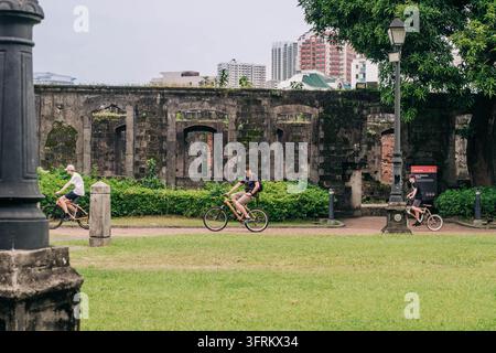 Manila, Philippinen, 23. September 2026. Touristen fahren mit Fahrrädern auf einem gepflasterten Weg vor einer alten Steinmauer mit gewölbten Öffnungen, Grünflächen und Mod Stockfoto