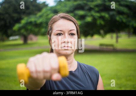Nahaufnahme-Porträt einer konzentrierten Frau, die ihren Arm nach vorne streckt, während sie eine gelbe Hantelstange während einer Outdoor-Widerstandstraining-Übung in einer Kneipe hebt Stockfoto