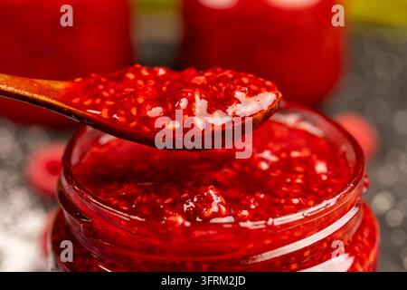 Rote hausgemachte geriebene Himbeeren in einem Löffel über einem Glas, frische geriebene Himbeeren in einem kleinen Löffel vor einem dunklen Hintergrund aus Schiefer mit Zucker Stockfoto