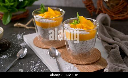 Zwei Gläser Chia-Pudding mit hellem Mangopüree und frischen Fruchtstücken. Die rustikale Szene bietet sanftes natürliches Licht auf einem dunklen s Stockfoto