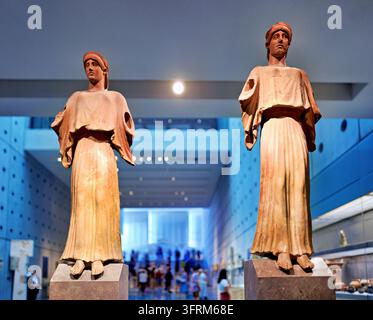 Zwei drapierte antike griechische Skulpturen von Frauen auf Steinpodesten in einer blau beleuchteten Museumsgalerie, mit verschwommenen Besuchern im Hintergrund, Griechenland Stockfoto