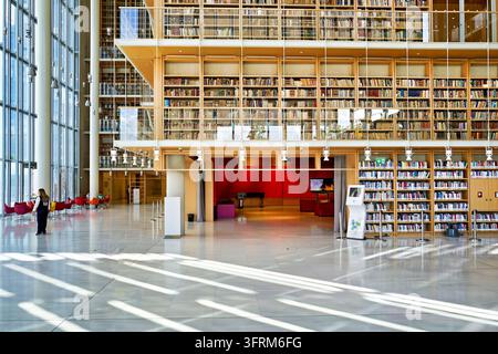 Holzbücherregale erheben sich hinter Glas im Atrium des Stavros Niarchos Foundation Cultural Center, während Sonnenlicht den Marmorboden streift. Stockfoto
