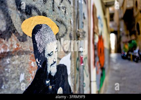 Aufgeklebtes Kunstwerk eines lächelnden bärtigen Mannes mit einem goldenen Heiligenschein auf einer mit Graffiti bedeckten Wand in einer schmalen gepflasterten Gasse in Neapel, Süditalien Stockfoto