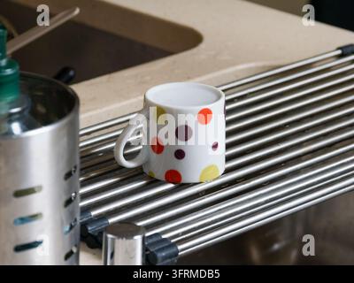 Weiße Keramiktasse mit bunten Punkten, die auf einem Metall-Trockenständer über einem Küchenwaschbecken steht. Stockfoto