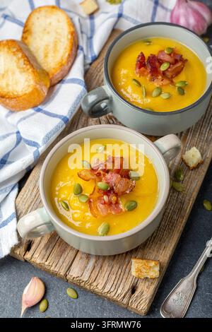 Hausgemachte Kürbissuppe mit knusprigem Speck und Croutons in Keramikschüsseln auf einem alten Holzbrett, selektiver Fokus Stockfoto