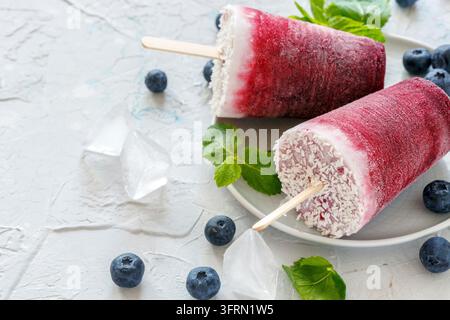 Hausgemachte vegane Beeren-Eis am Stiel mit Kokosmilch, frischen Blaubeeren und grüner Minze auf einem weißen Betonstisch Stockfoto