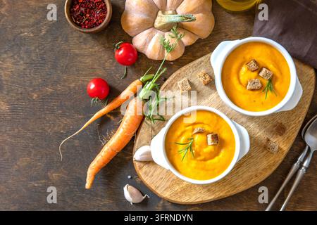 Kremige Kürbissuppe in weißen Schüsseln, garniert mit gerösteten Croutons und frischen Kräutern, arrangiert mit Karotten, Kirschtomaten, Gewürzen und rustikalem Holzservierlöffel Stockfoto