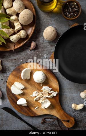 Ansicht von oben über frische Pilze, Knoblauchzehen, Olivenöl, Pfefferkörnern und einer schwarzen Pfanne, die auf einem rustikalen Tisch mit einem Holzschneidebrett angeordnet sind für Stockfoto
