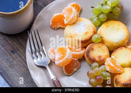 Frisch gebackene ukrainische Syrniki auf einem Keramteller mit geschälten Mandarinenstücken, grünen Trauben und einer Tasse Kaffee auf einem rustikalen Holztisch. Stockfoto