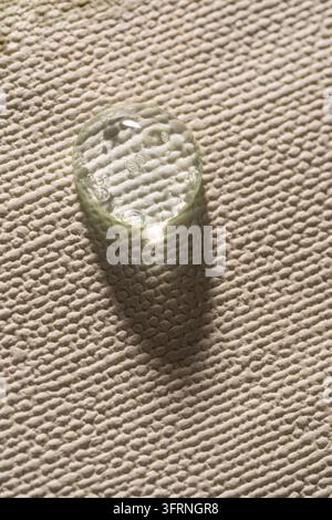 Ein transparenter Geltropfen ruht auf texturierter weißer Leinwand, fängt weiches Sonnenlicht ein und wirft einen langen Schatten. Die Nahaufnahme betont glänzende Reflexionen, b Stockfoto