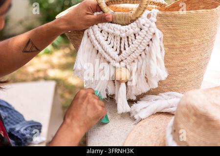 Nahaufnahme eines Handwerkers, der eine handgefertigte Makrame-Dekoration mit weißem Baumwollgarn, Quaste und Holzperle an einen gewebten Korb während eines Outdoor-Handwerks anbringt Stockfoto