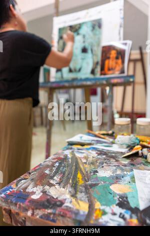 Studentin malt auf einer Leinwand während eines Malunterrichts in einem Kunststudio, umgeben von Pinseln, Farbtuben, Gläsern, Paletten und farbenfroher Kreativität Stockfoto